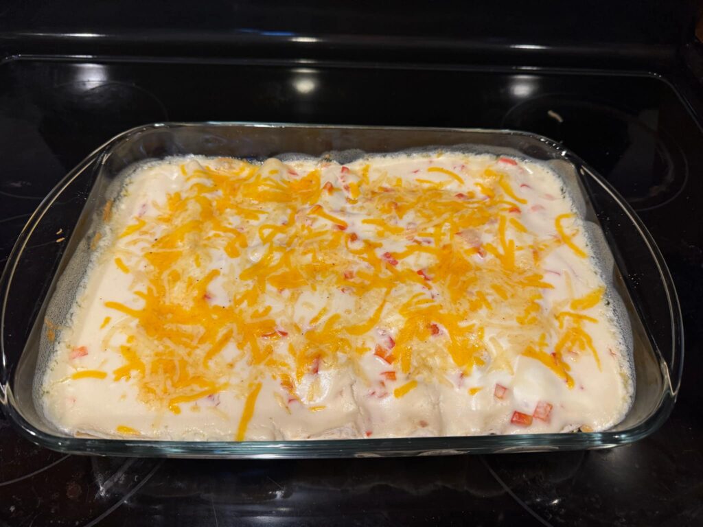 A glass baking dish filled with creamy chicken enchiladas, topped with melted cheddar cheese and diced red bell peppers.