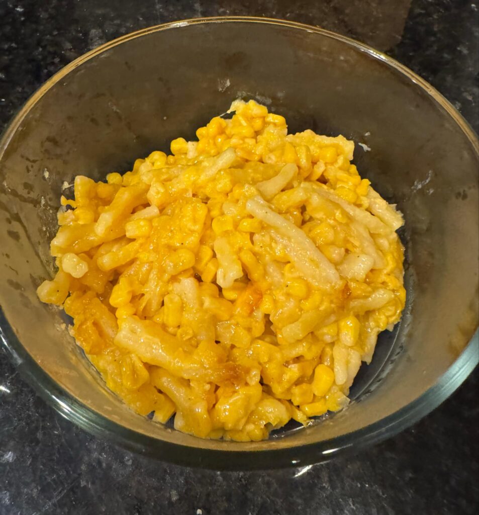 A bowl of cheesy corn casserole with pasta, featuring a golden, gooey texture from melted cheddar and sweet corn kernels.