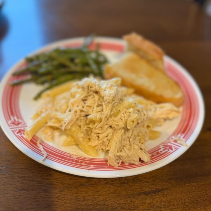 A plate of creamy Olive Garden Chicken Pasta with shredded chicken and penne, served alongside roasted green beans and garlic bread.