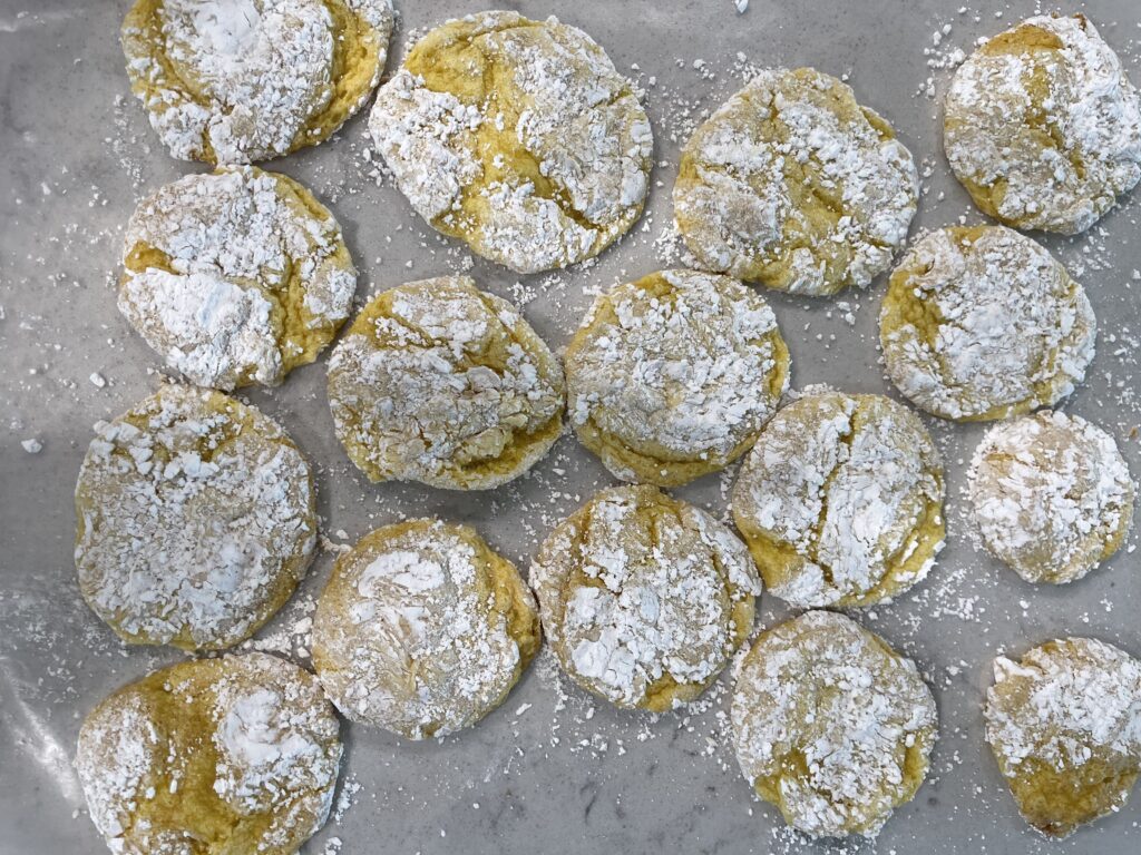 A batch of freshly baked lemon crinkle cookies with a golden yellow hue, coated in powdered sugar, resting on a parchment-lined baking sheet.
